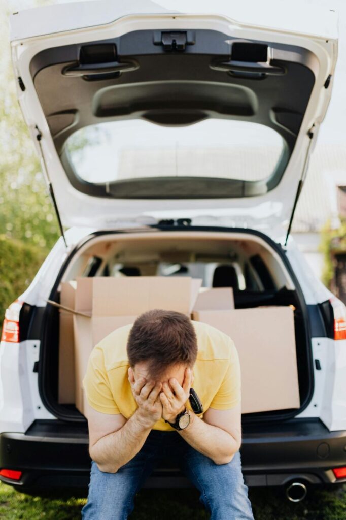 A man sits on the back of his car's open hatchback filled with moving boxes, his head in his hands in a gesture of stress. Easing the stress of moving can help tremendously when learning how to make a house feel like a home