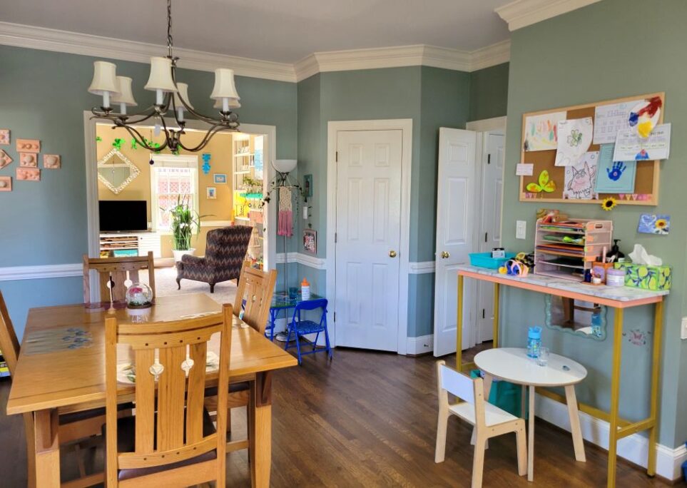 Breakfast area of kitchen facing the closed pantry door (from standing near the end of the counter island). To left is kitchen table and chairs. To right is short wall with console table and round white toddler table. To right of closed pantry door are the open doors leading into the foyer. To left of pantry door is second toddler table where kindergartener prefers to sit, along with a floor lamp in corner with macrame design hanging from switch and some artwork on the small wall space above table. To immediate left is wide opening into the back living room.