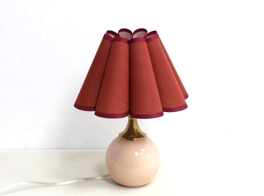 A lamp with a rust red scallop shaped cone shade with maroon trim on the top and bottom, and a cream coloured round ceramic base with gold-coloured neck