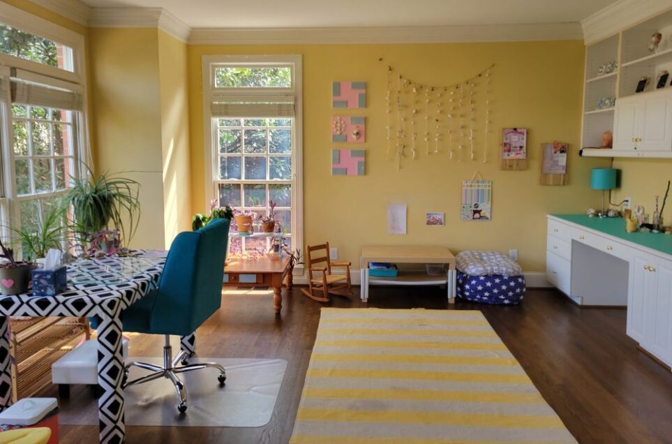 View of study from standing in double French door entryway, facing back wall with a coffee table flanked by child rocking chair on left side and star-spangled beanbag chair filled with blankets and a pillow on top on right side. On wall above hangs seashells strung along ribbons hanging from bell strand near top of ceiling, three painted canvases with tiles and shells, and other collages. To left along wall is built-in desk and shelving. On left side of back wall is window with another coffee table with plants on top. Along left wall are more windows and, in front, more plants as well as free-standing desk and chair.