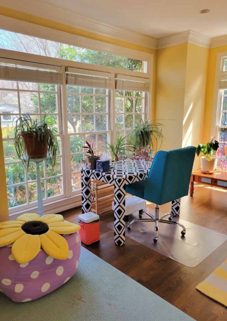View of study facing corner of back wall with east-facing windows and free-standing desk and chair. To near left is periwinkle-with-white-polka dots beanbag case filled with pillows and a bright flat sunflower baby pillow on top for lounging (a pale blue flat area rug lies underneath in that corner). Behind it in front of window is blue-painted wooden plant stand supporting airplane plant. To right and facing windows is black and white patterned upholstered desk with teal swivel chair, and plants on top and in front of it. Toward far right in front of window is another airplane plant on a plant stand. In far right corner by partially-seen south-facing window is a coffee table with more plants.