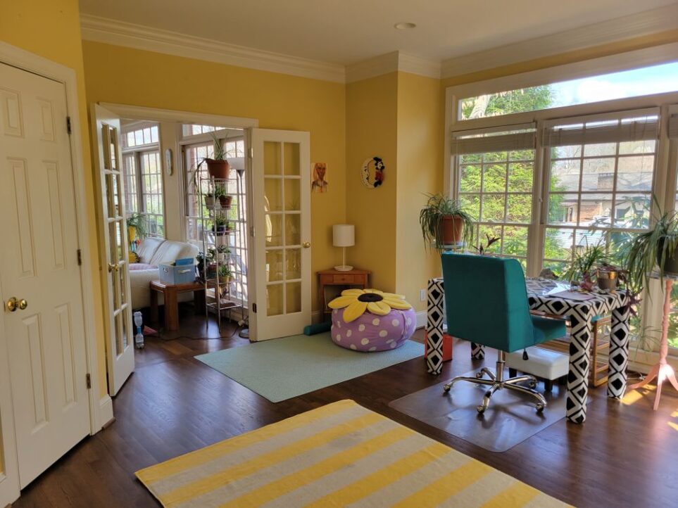 View of study from the back corner of built-in desk, facing the cosy corner with blue rug and open French doors to its left. A yellow and white striped rug spans the center of the floor, partially-seen below. To right, facing the windowed-wall, is free-standing desk with chair and several plants on and around it in front of windows. Straight ahead is cosy corner with fabric beanbag case filled with pillows and topped with yellow sunflower pillow. A small corner table and white lamp are in the corner. A painting and moon decoration hang on the wall above. To the left are the open French doors leading into the sunroom. To the far left is the closed study closet door.
