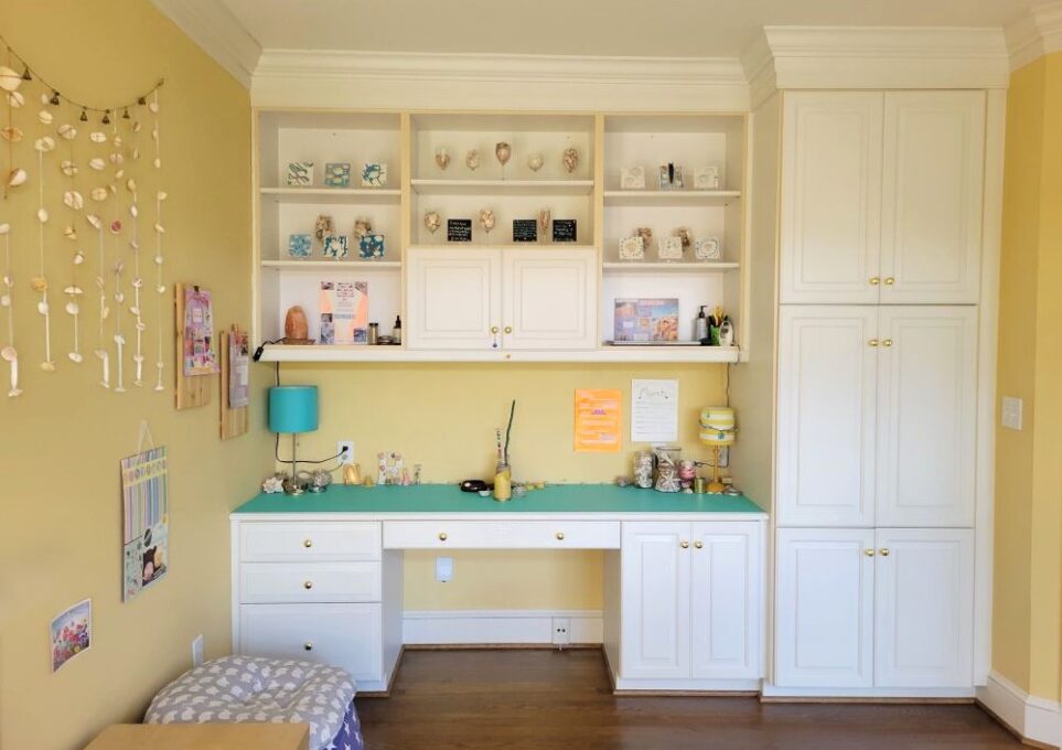View of study's built-in desk and surrounding cabinets and shelves. Aqua-coloured peel-and-stick wallpaper lines the desktop, and a small table lamp is in each corner, surrounded by seashells and other meaningful decor. On shelves above are decorations - shells in wine glasses, painted tiles with seashell art, a salt lamp, etc. Hanging on wall to left are collages and seashell ribbon strands. All of the surrounding beauty and playfulness makes the study one of my favourite creative spaces.