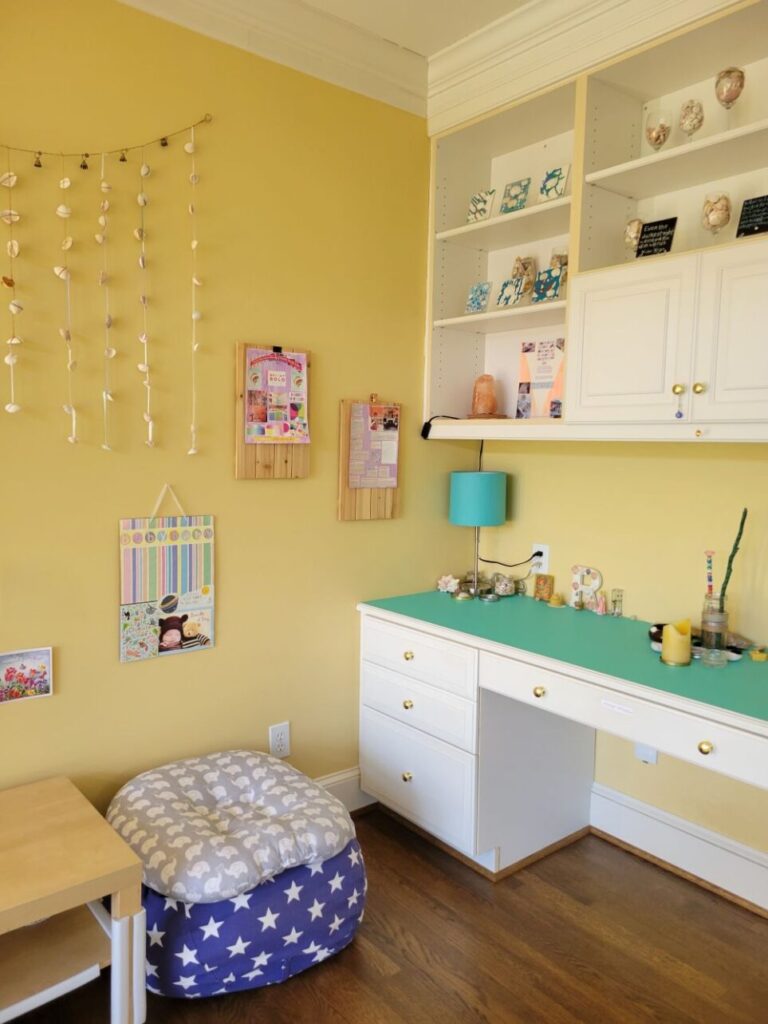 Corner of study with built-in desk and shelving filled with homemade decorations (painted tiles with shell art, shells in wine glasses, etc.). Covering built-in desktop is aqua-coloured peel-and-stick wallpaper. A blue lamp stands in corner among keepsakes and seashells. Other meaningful decor and items are along length of desk toward wall. To left is a partially-seen coffee table for children's art and crafts; to its right a blue beanbag case with white stars, filled with old blankets, and a baby pillow on top for a seat. Homemade decor and collages hang on the wall above it.