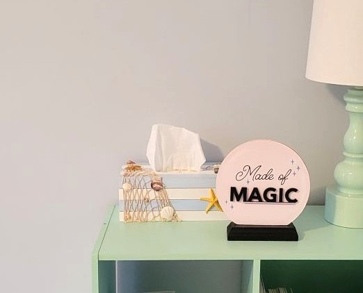 Nautical-themed wooden tissue box holder with light blue and white horizontal shiplap stripes, yellow sea star, and fish net with shells in it, sitting on sea green cubby shelving unit next to sea green touch lamp with white shade and decorative placard that says 'Made of Magic'
