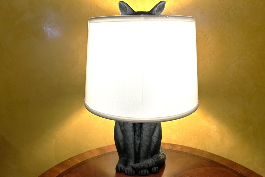 A plain light lampshade with the light turned on hides a base in the shape of a cat. Its lower body (legs and tail) are seen standing below the shade, and its ears stick out through the top of the shade.