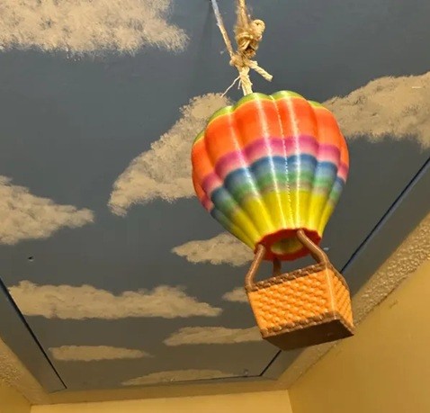 A plastic 3D-printed rainbow coloured hot air balloon hangs from a cloud-painted blue ceiling