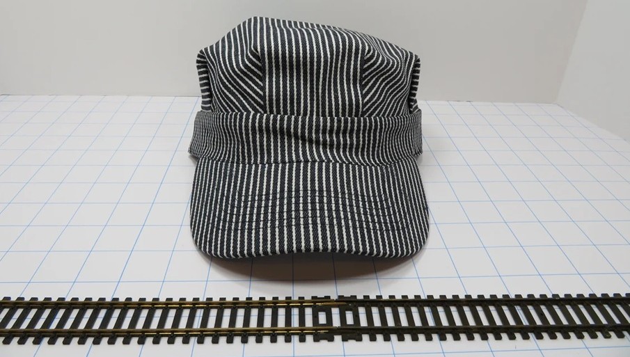 A hickory-striped train engineer's hat rests on a surface with a stretch of toy train tracks in front of it