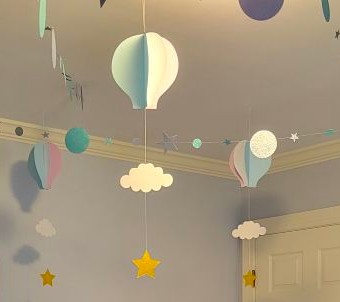 Blue and pink paper hot air balloons with white paper cloud cutouts and gold paper star cutouts hanging on the string below, with a garland of paper cutout blue stars and circles strung horizontally between balloons