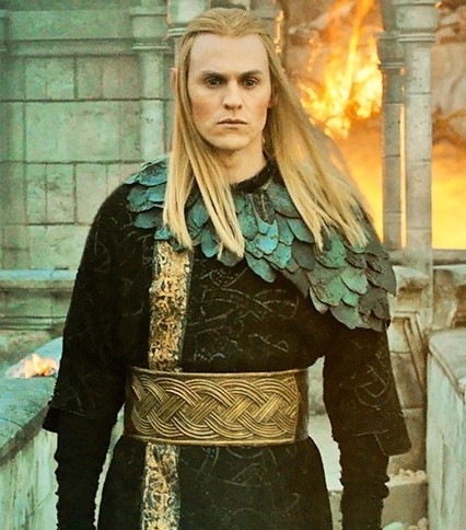Image of Charlie Vickers portraying Annatar in season 2 of The Rings of Power. Wearing elegant armor and with a serious expression, he stands on a balcony in Eregion while fires rage in the background from battle shots