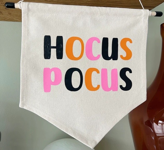 Close-up photo of an off-white cloth pennant banner hanging with the words 'Hocus Pocus' in black, orange and pink