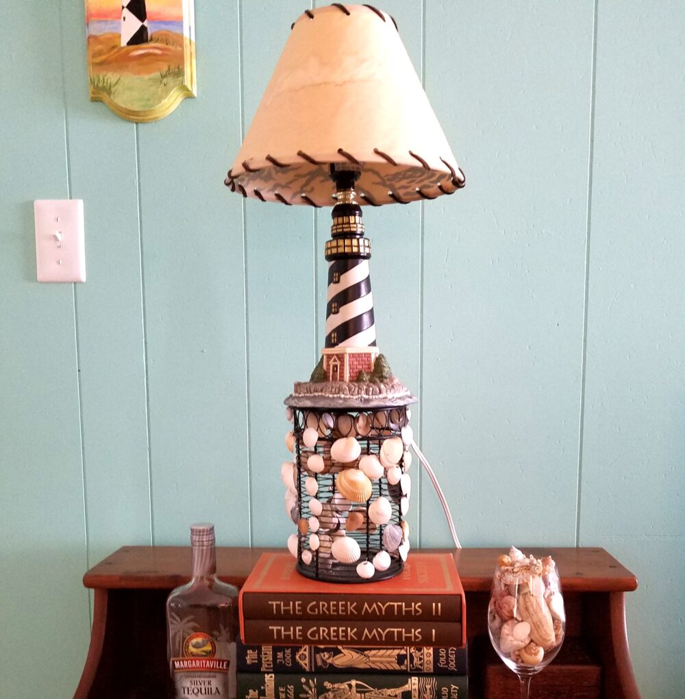 A small lighthouse-shaped lamp is elevated on a small brown end table atop a seashell-adorned upturned cylindrical metal wire container that stands atop a stack of old tomes (The Greek Myths, The Ancient Persians, etc.)