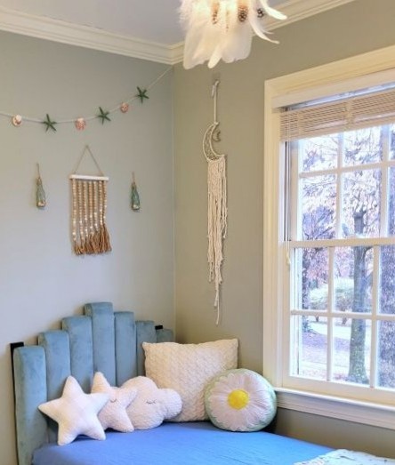 Corner of boy's bedroom has upholstered blue headboard with several soft throw pillows in corner for a reading nook next to the window, and several calm decorations hanging on wall above including macrame crescent moon-shaped dream catcher, strand of seashells and starfish, and wooden bead decoration