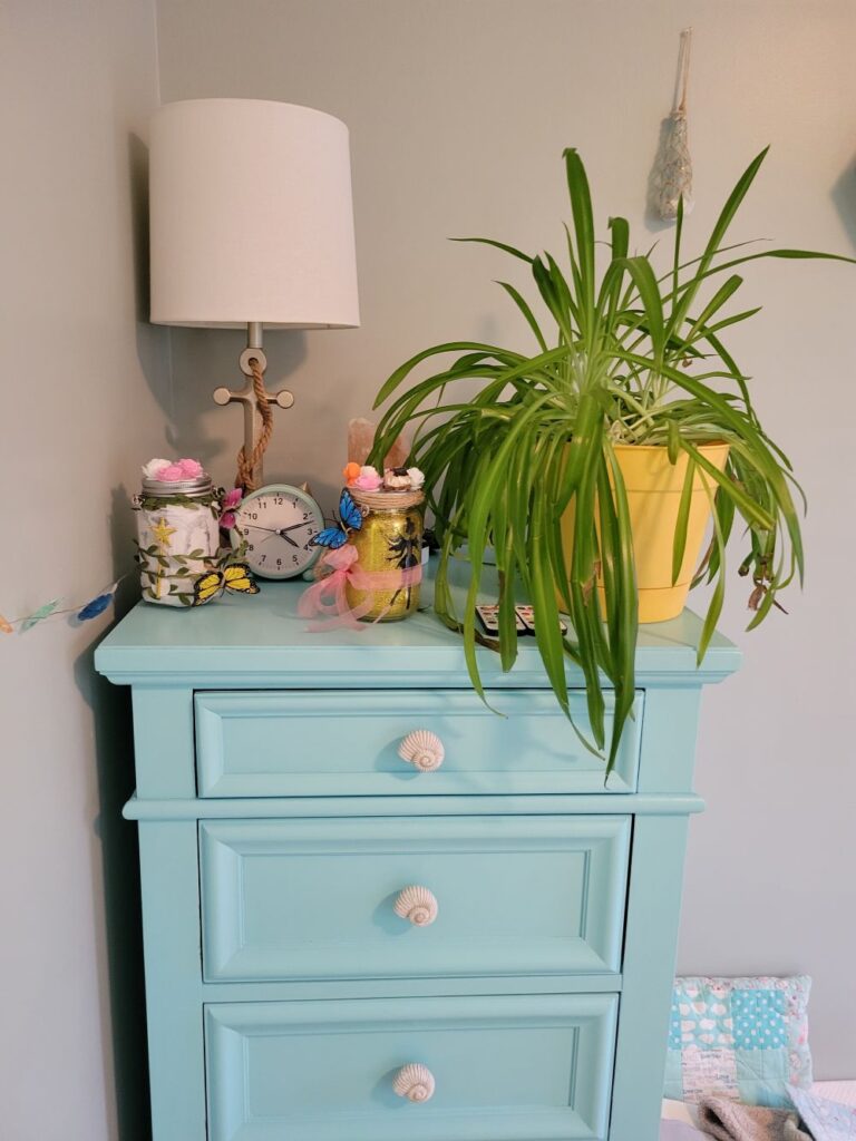 The corner of the boys' bedroom features a light blue-painted dresser with spiral seashell drawer knobs, and an anchor-shaped lamp on top next to an airplane plant, an analog clock, and two decorative fairy lanterns