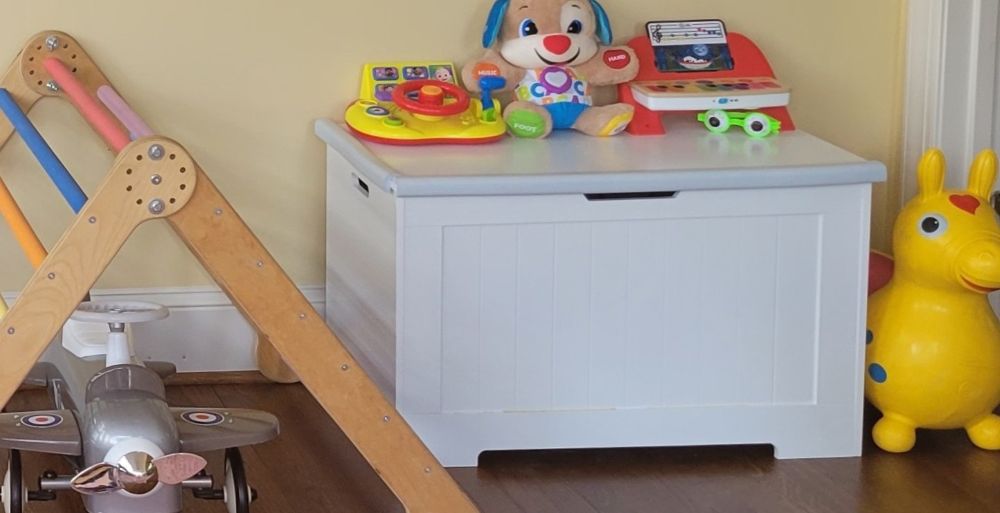 A handsome white wooden toy box next to a Pikler triangle with a ride-on airplane underneath on the left, and a ride-on inflatable mini horse to the right. A few toys sit on top of the toy box as well. Along with a toy display shelf, use a toy box like this to keep toys neatly off of the floor in an attractive spot