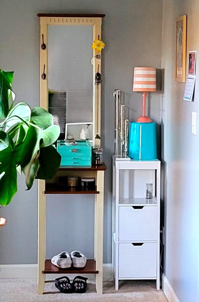 In a corner of the primary bedroom is a narrow square white table with an upside-down blue trash bin on top and a small peach-and-white striped table lamp on top of the upturned bin, for elevation. Next to the bin/lamp on the white table is a tall necklace holder, and next to the table is a narrow standing vanity with a tall mirror and a blue jewelry box on top. On the wall in the corner are a couple of artworks, for a pleasant and airy corner