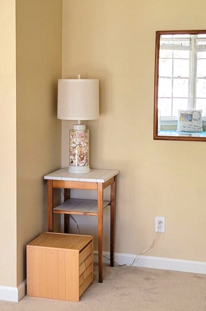 An odd corner in the upstairs office/bedroom features a square end table (covered in shiplap peel-and-stick wallpaper on top) with a large lamp on top filled with seashells. To the right on the wall is a mirror to reflect light from the windows on the opposite wall, and on the floor next to the small end table in the very small corner nook is a small square wooden box filled with drawers of wooden building blocks for children to play