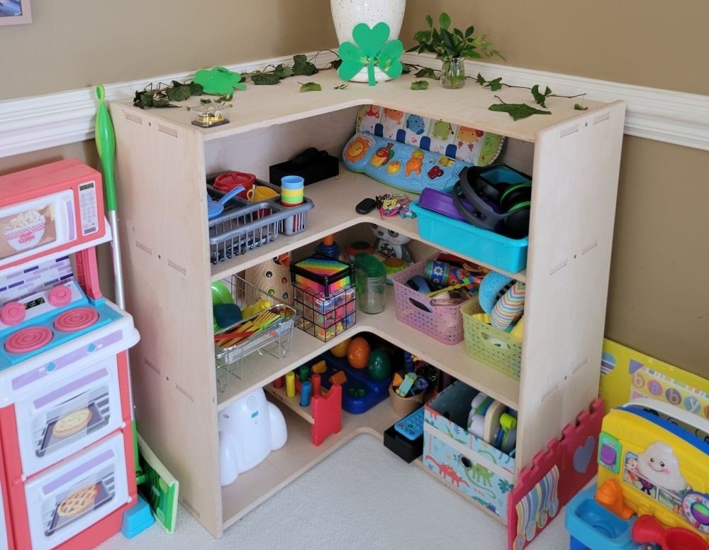 Montessori corner toy display shelf filled with organized toys on the three shelves, and decorated with ivy vines on top (along with green cut-out shamrocks for St. Patrick's Day)