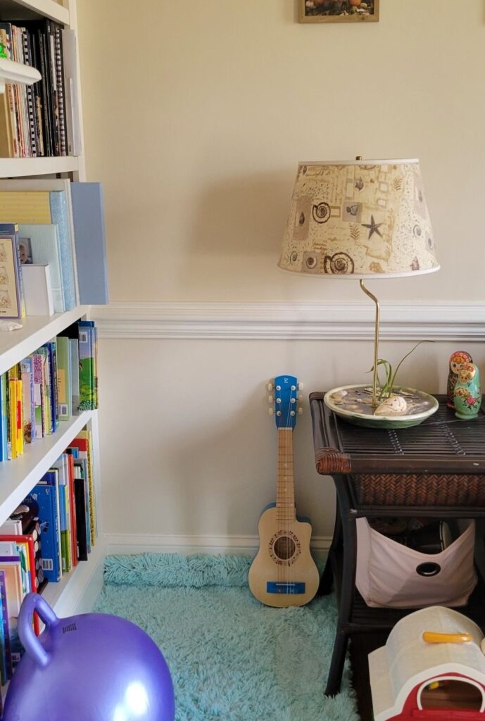 This cosy corner in the front living room features an a small aqua shag rug overtop a bath mat for comfortable seating, a built-in bookcase on one side with children's books on the lower two shelves, and a square dark brown wicker end table next to the rug with a large seashell lamp on top for reading light, and a couple of Matryoshka dolls on top. It's the perfect nook for reading with children. A large purple bouncy ball is docked against the bookcase, and a few toys like a small toy guitar, and barn are nearby