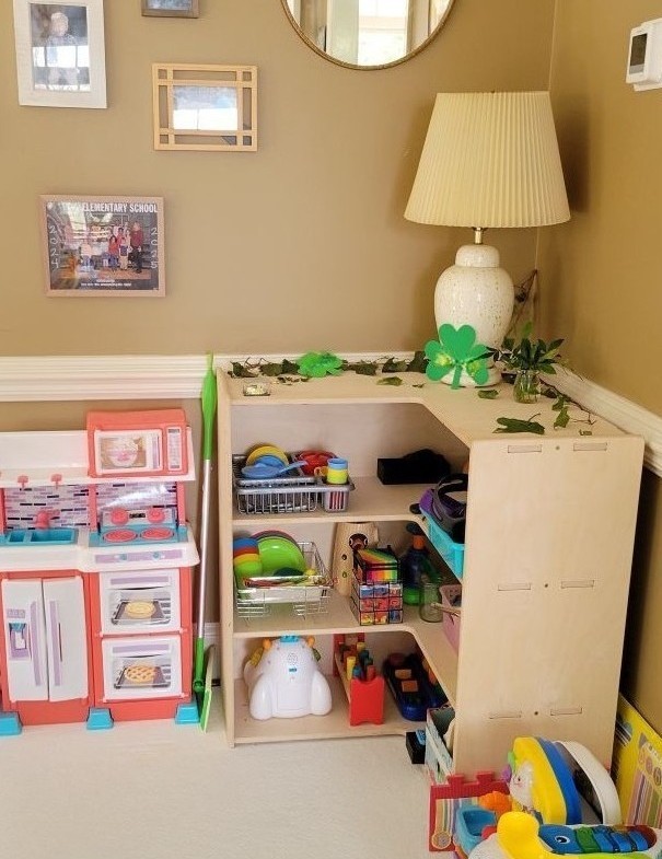 Corner toy shelf in living room with lamp on top in corner alongside decorations. Mirror and framed photos hang on wall above and toys are neatly arranged on shelves below lamp