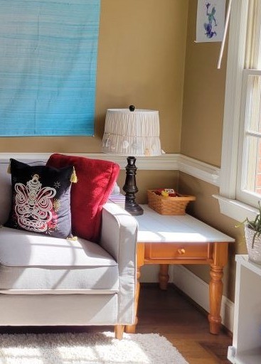 The corner of the back living room is filled with an end table with a lamp on top and tissue box, next to one end of the couch with some throw pillows below a thin fabric blue tapestry hanging on the wall and a blue/purple print hanging on other wall next to the window that provides natural light as well