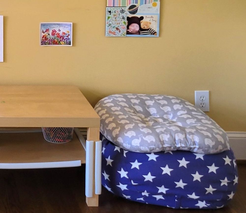 A plush animal storage bag (blue with white stars) filled with blankets and with a cushion on top sits next to a coffee table and serves as a spot for a toddler to sit and colour at the table
