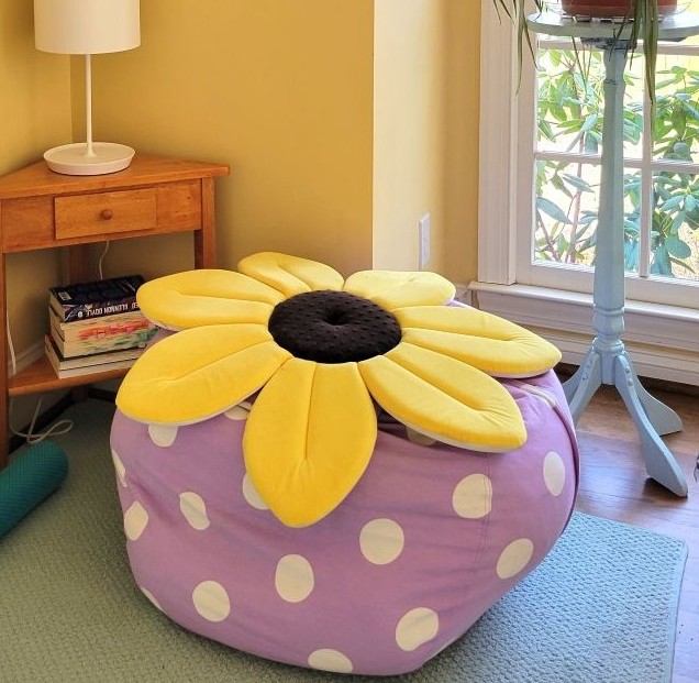 A plush animal storage bag (periwinkle with white polka dots) filled with blankets and pillows and with a large flat soft yellow flower cushion on top sits next to a small wooden corner table with a white stick lamp on top, as well as a light blue-painted plant stand with an airplane plant on top (barely in view). The plush animal storage bag serves as a lounging spot to sit and read