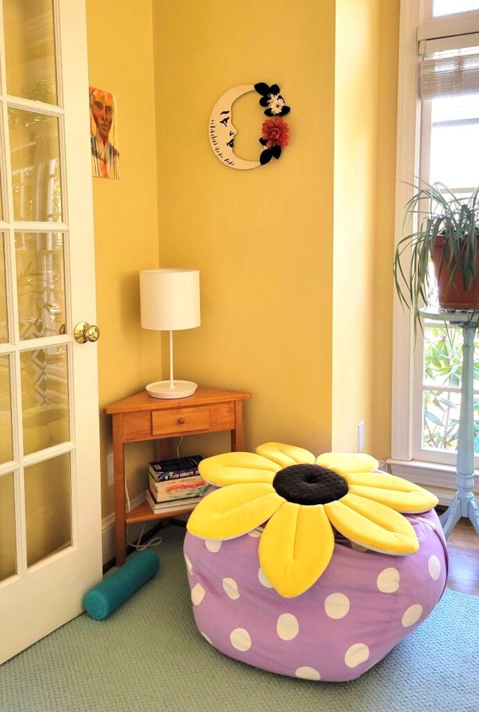 Create inviting corners like the one shown in this photo featuring a small triangular wooden corner table with a white stick lamp on top, a large plush animal case filled with pillows and blankets for a lounging cushion with a soft flower-shaped flat pillow on top, and pretty decor hanging on the wall above the corner table and lamp. A small blue rug fills the corner and several books are stacked on the lower shelf of the corner table. Nearby is a window with a plant on a plant stand in front of it
