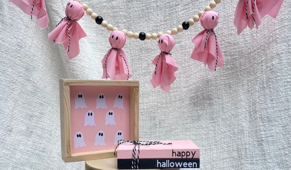 Photo of a pink ghost garland hanging across top of frame, a wood framed mini minimalist painting with pink background and white ghosts resting atop of wood stool, and a wooden book stack with a black wood 'book' under a pink wood 'book' next to it, pink with 'happy' and black with 'halloween' on the spines