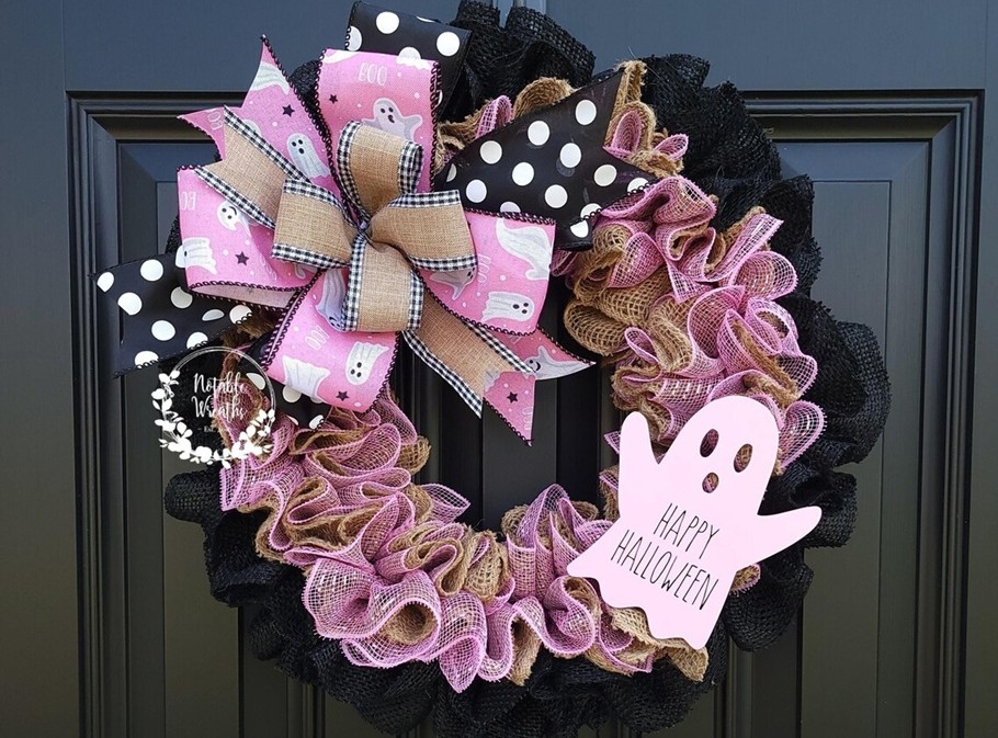 A pink and black Halloween wreath hangs on a black-painted door. It's adorned with a pink ghost that says 'Happy Halloween', burlap, and pink and black bows with various patterns.