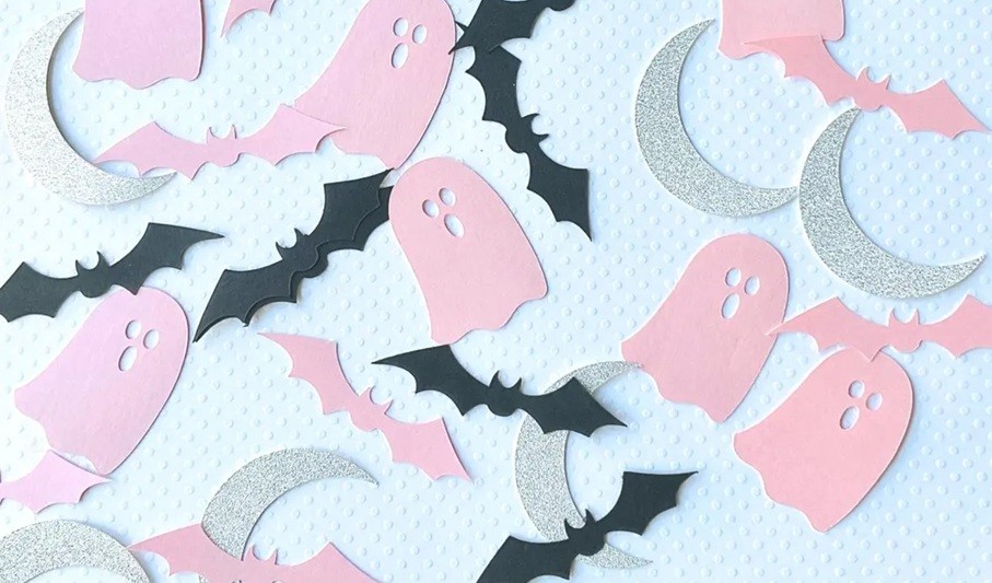 Close-up photo of Halloween confetti spread over a white surface with pink ghosts, pink and black bats, and glittery silver crescent moons