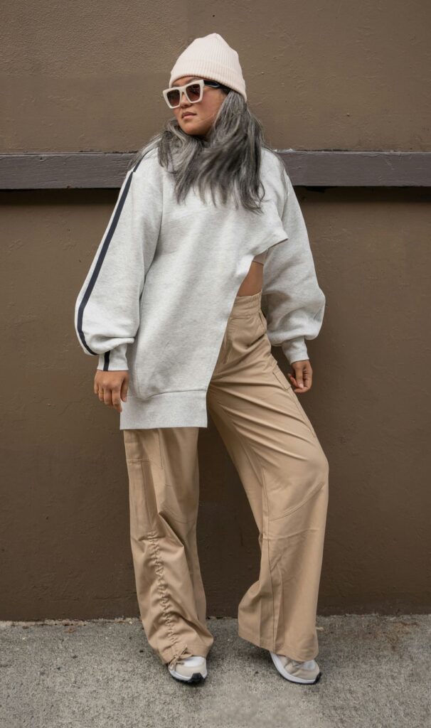 Woman posing in asymmetrical light gray sweatshirt with cream knit beanie, pale pink-rimmed sunglasses, and khaki cargo pants with sneakers