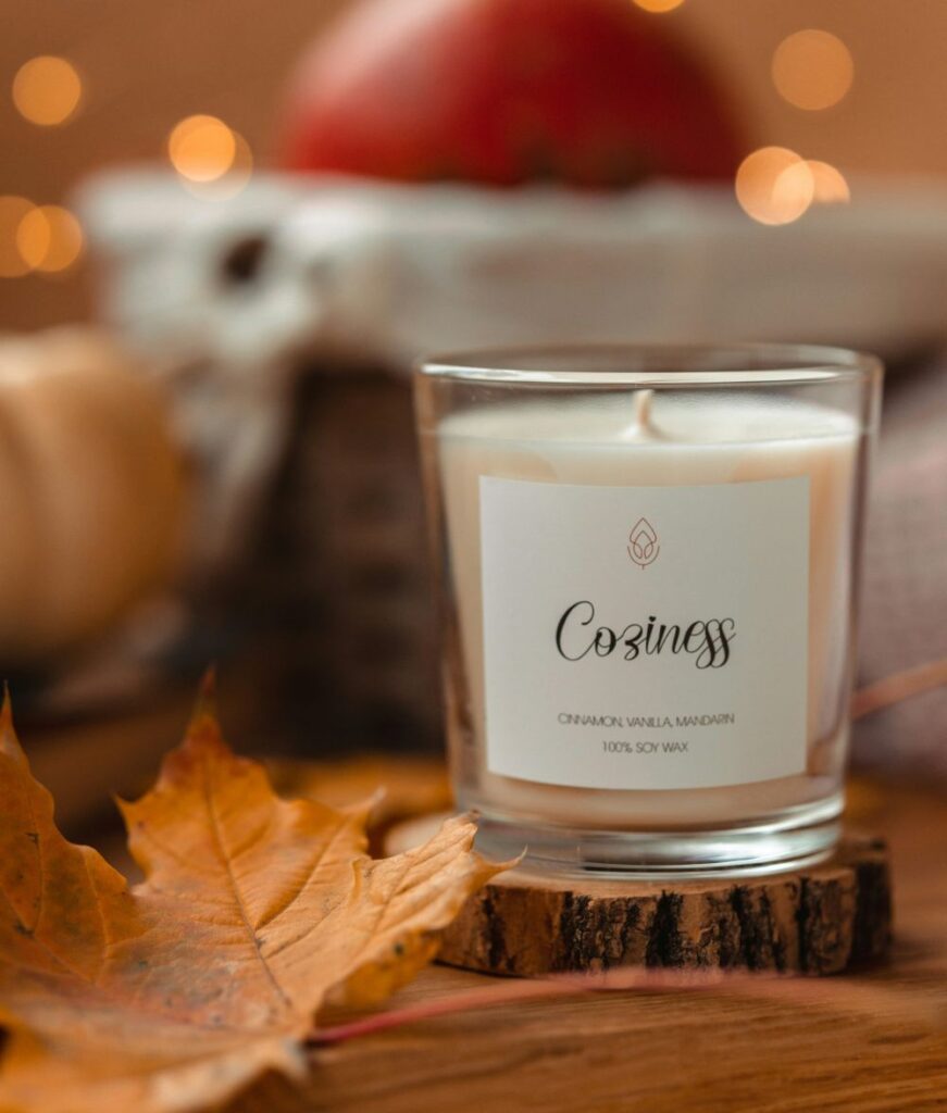 A candle poured in a glass jar called 'coziness' sits on a wooden tree limb cross-section coaster next to a brown maple leaf in an autumn vignette. Its label reads 'cinnamon vanilla mandarin' and '100% soy wax' for autumn-themed clean burning candles