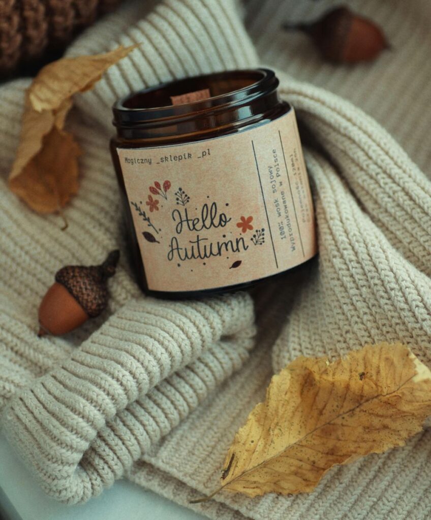 A woodwick candle in a brown glass jar with a label titled 'Hello Autumn' and '100% wosk sojowy' (Polish for soy wax) rests in the nook of a cream-coloured folded sweater amidst acorns and brown dried leaves (Варшава, Мазовецьке, Польща)