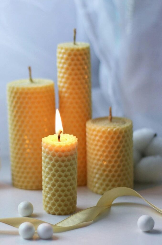 A photo of clean burning candles: four honeycomb-indented beeswax pillar candles of different heights (with the shortest one lit) amidst small white balls and a gold ribbon in front of a white backdrop in Пермь, Пермский край, Россия