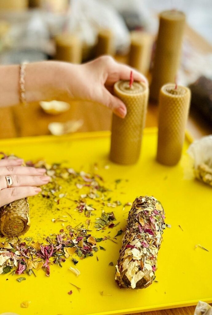 A woman's hands are seen making beeswax pillar candles by rolling the wax longways on a table with scattered dried flowers and herbs, a beautiful and creative process for make scented clean burning candles (Cherkasy, Cherkas'ka oblast, Ukraine)