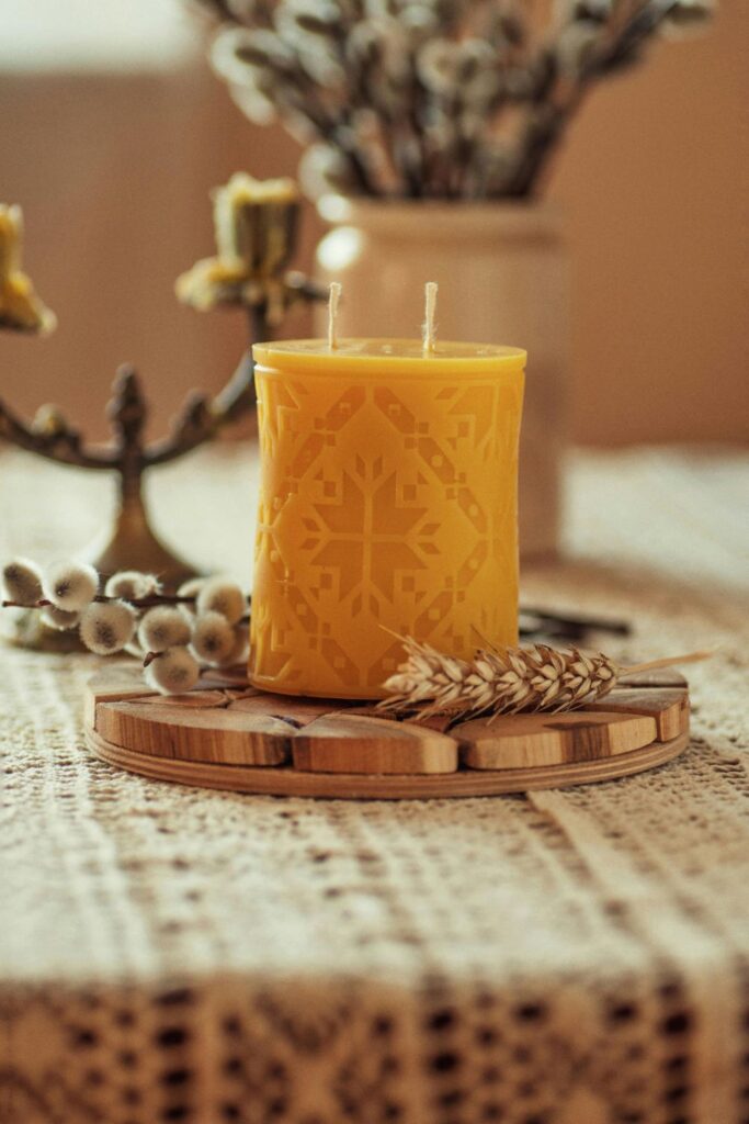 A beeswax pillar candle beautifully carved with a geometric pattern rests atop a round wooden trivet on a table covered in a cream lace tablecloth and other neutral-coloured decor in Варшава, Мазовецьке, Польща