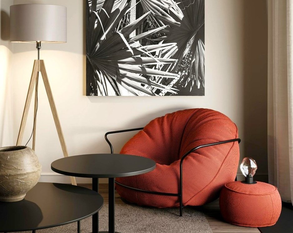 The corner of a living space with a large rust-red beanbag chair and matching small ottoman next to it, small black table, large black and white art print on the wall, and a floor lamp