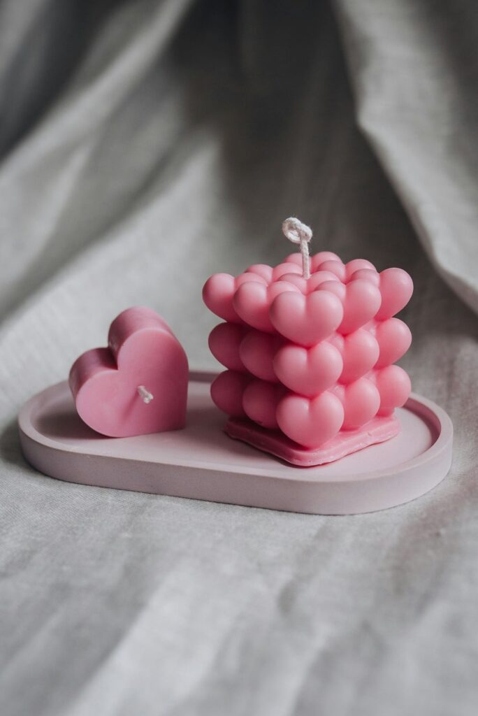 Photo of two pink clean burning candles made of soy wax: one is smaller and heart-shaped and rests on its side on a small ovular tray, and the other next to it on the tray is shaped like a cube composed of many small hearts (three to a row/column)