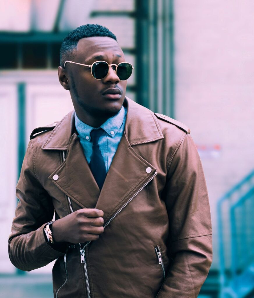 A man wearing sunglasses and a brown leather jacket over a blue button-down collared shirt with navy tie poses in New York City, where fashion is important to everyone!
