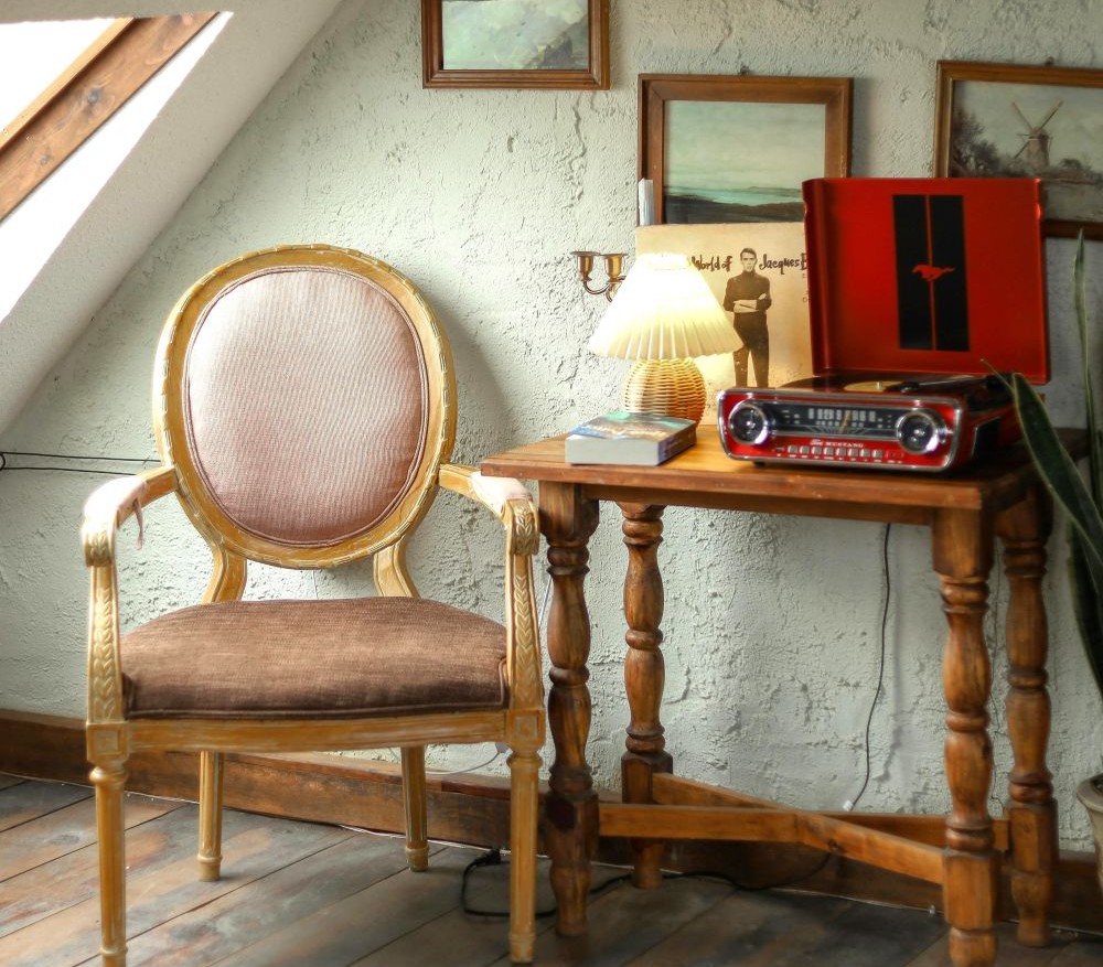 The corner of an attic with a low slanted ceiling and window/skylight next to an upholstered chair and small wooden table with a small wicker table lamp and other decor on top, and framed art on the stucco wall behind it