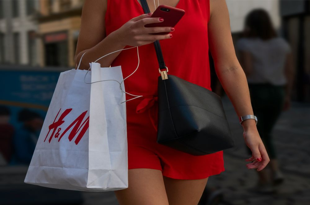 Close-up photo of a woman in a red outfit walking and looking at her phone whist carrying a black purse over one shoulder and an H&M shopping bag on her arm. H&M has been criticized for unethical labour conditions in various parts of its supply chain as well as lack of transparency, even after its public statements claiming commitment to fair labor standards
