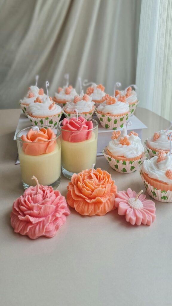 Several peach-coloured cupcake-shaped candles with white 'icing' and small peach flower embellishments are arranged next to a couple of 'flower pudding candles' in clear glass jars that resemble pale yellow pudding with pink and orange flowers on top, and a couple of carved flower candles in pink and orange