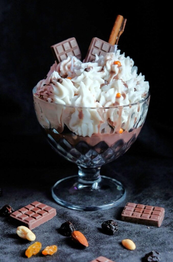 A clear glass dessert bowl on a stem holds a candle that resembles a delicious chocolate puddindg dessert with whipped cream and pieces of chocolate bar sticking out of it (Bucureşti, România)