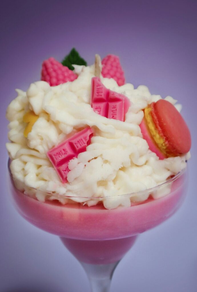 Close-up photo of a clear dessert glass filled with a candle that resembles a raspberry sorbet dessert topped with white whipped cream, tiny macarons, raspberries, and broken chunks of pink milk chocolate bar