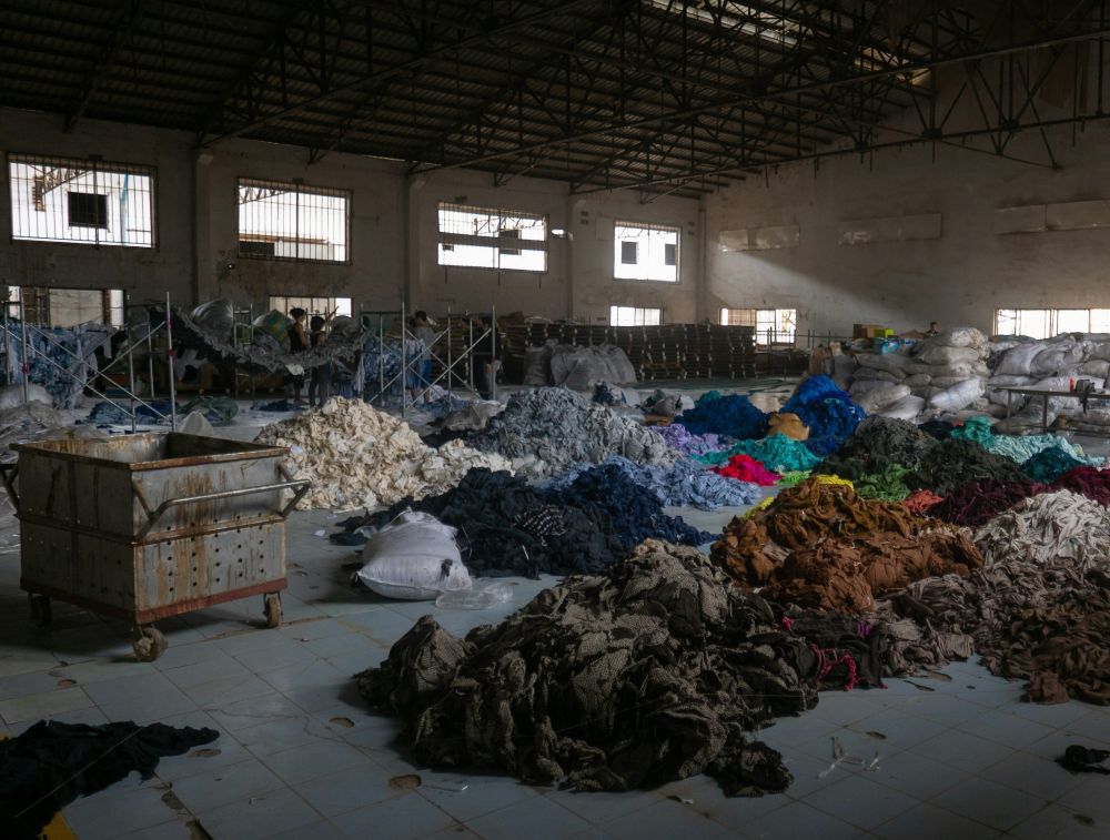 Dismal photo of a large abandoned textile factory in Phnom Penh, Cambodia with messy mounds of dirty fabric dyed in various colours and a large rusty metal bin
