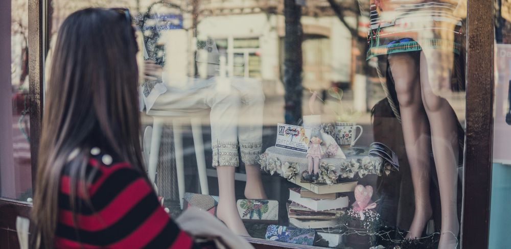 A woman in a red and black striped shirt stands outside a shop display window and looks through at the various decorations and clothing on display on two mannequins. Fashion is important because it provides us a creative outlet for showcasing our personal appearance