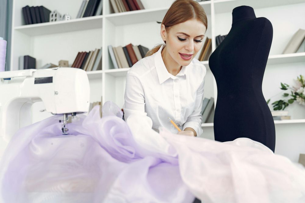 A woman in a white blouse writes notes with a pencil next to a black mannequin in front of a sewing machine with light periwinkle and lavender tulle fabric swaths