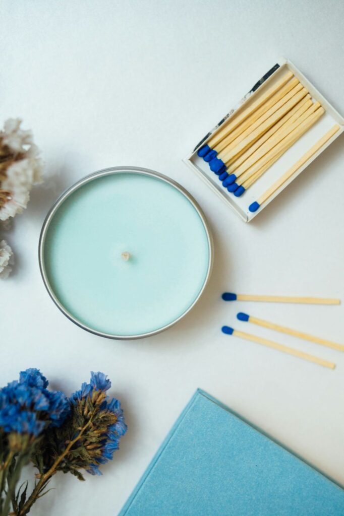Top-down photo of a candle with light blue-coloured soy wax poured in a round metal tin, with a box full of blue-tipped matches next to it, and a blue book and some blue flowers nearby