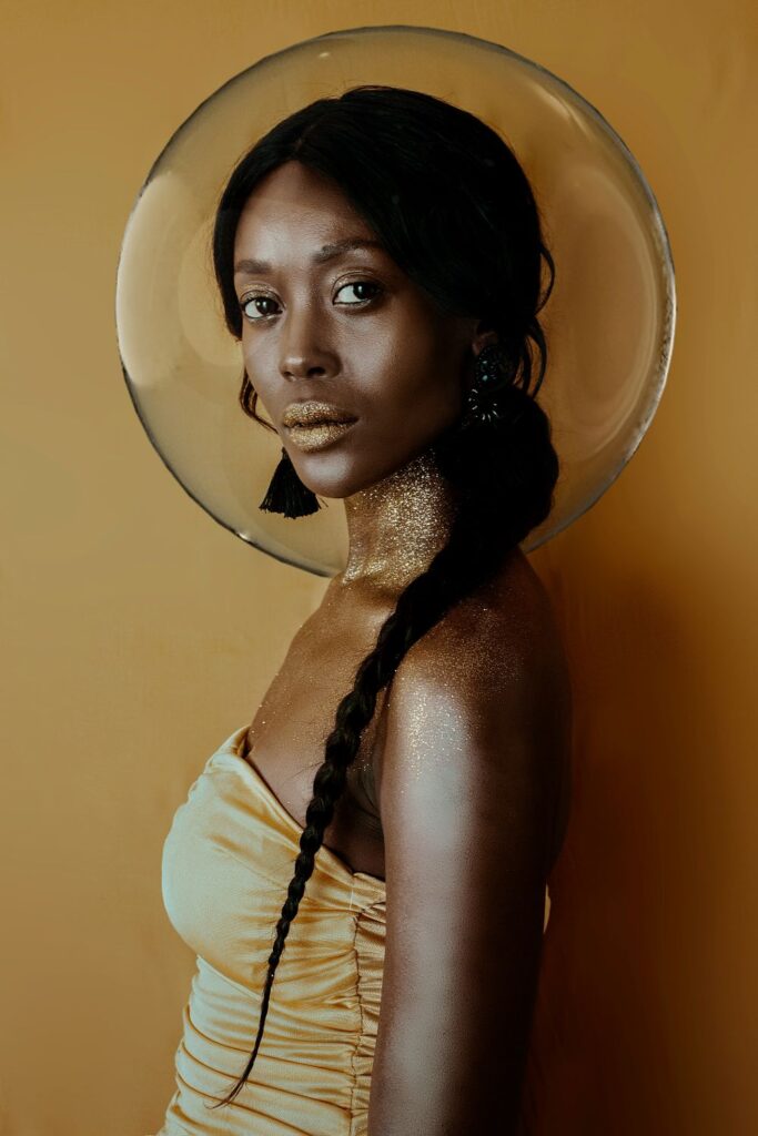 Profile portrait of a model wearing a sleeveless gold shirt with bunched fabric and gold glitter on her neck, shoulders and lips, facing the camera, with a large clear bubble behind her head (which appears to surround her head)